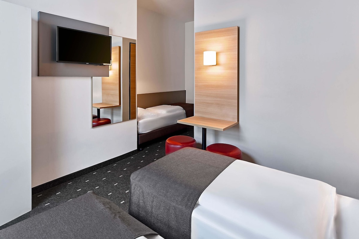 A modern hotel room with a bed, a TV mounted on the wall, a bedside table, and a small seating area with a red stool.
