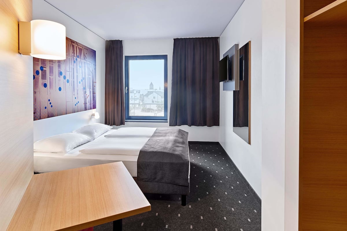 A hotel room with a bed, a small table, a bench, a TV, and a window with curtains.