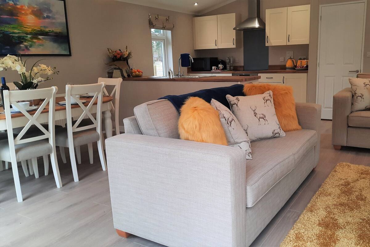 A modern living room with a grey sofa, a dining area with white chairs, and a kitchen in the background.