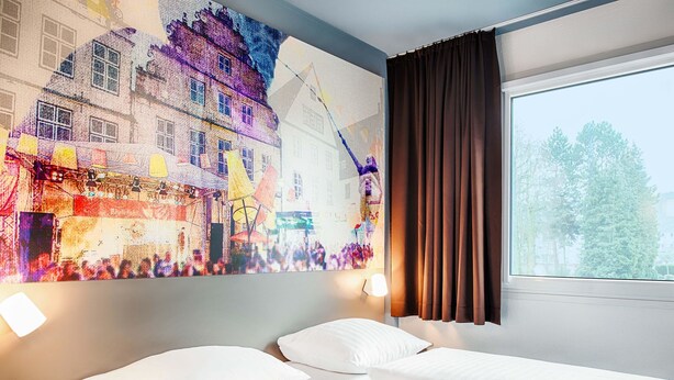 A hotel room with two beds, a large window with curtains, a wall-mounted lamp, and a colorful mural. - B&B Hotel Bielefeld-Ost (Bielefeld)