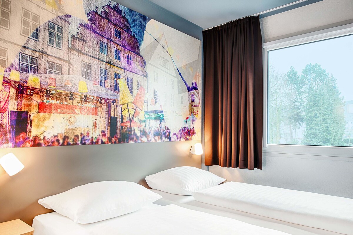 Habitación de hotel con dos camas, un ventanal con cortinas, una lámpara de pared y un mural colorido.