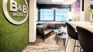 A B&B hotel lobby with a green wall, a reception desk, a seating area with a sofa and chairs, and a large window offering a view. - B&B Hotel Bielefeld-Ost (Bielefeld)