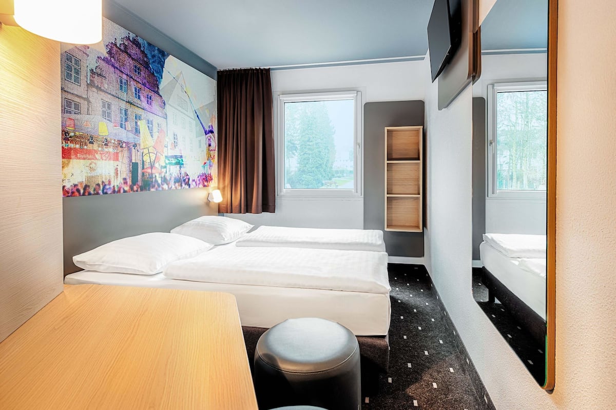 A hotel room with two beds, a desk, a chair, a wall-mounted TV, and a window with a view of trees.