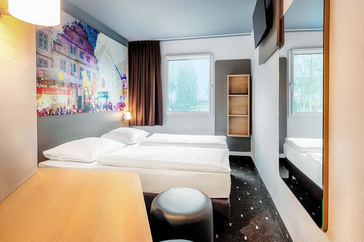 Habitación de hotel con dos camas, un escritorio, una silla, un televisor montado en la pared y una ventana con vistas a árboles.
