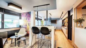 A modern kitchen with a dining area, a TV mounted on the wall, and a window with a city view.