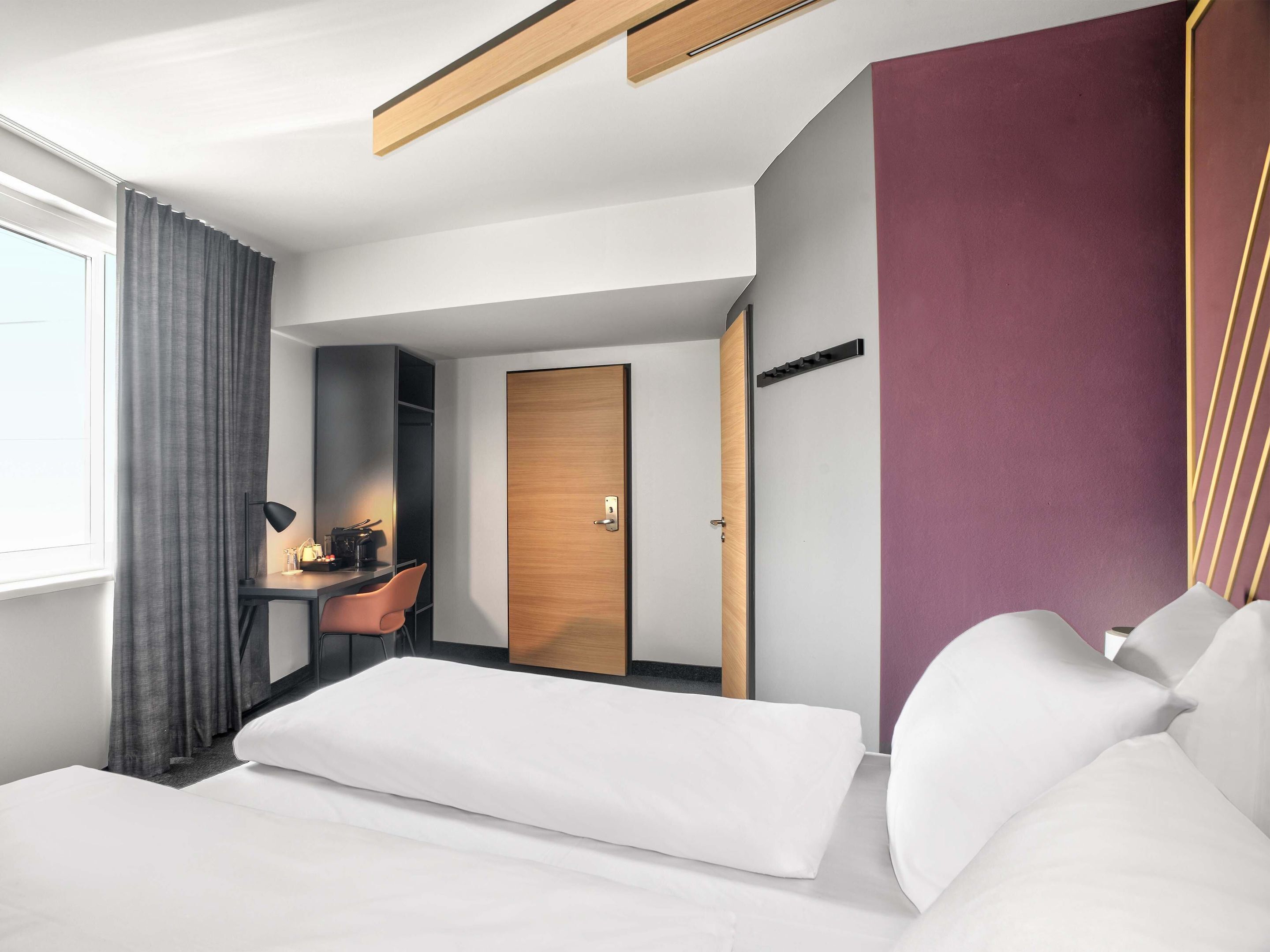 Una habitación de hotel moderna con cama, escritorio con silla, armario y un amplio ventanal con cortinas.