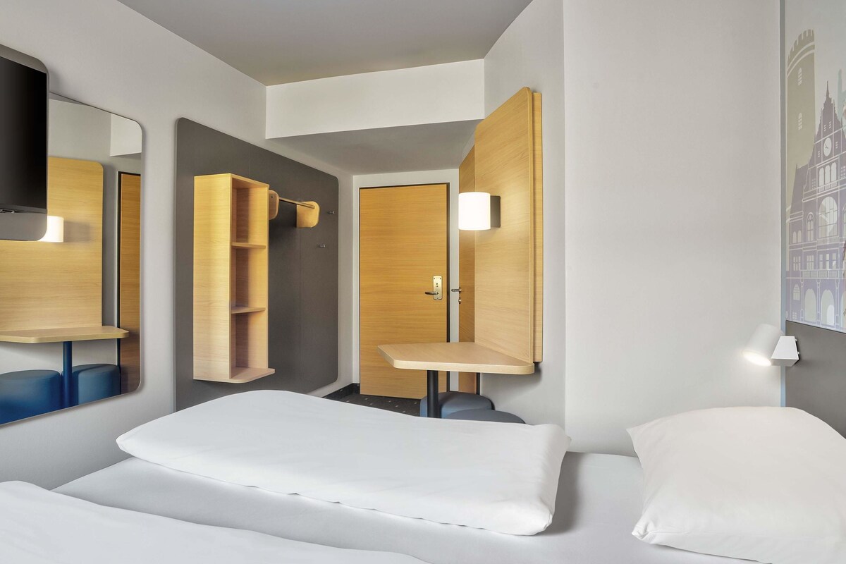 Ein modernes Hotelzimmer mit Bett, Schreibtisch, Spiegel und einer Wandablage.