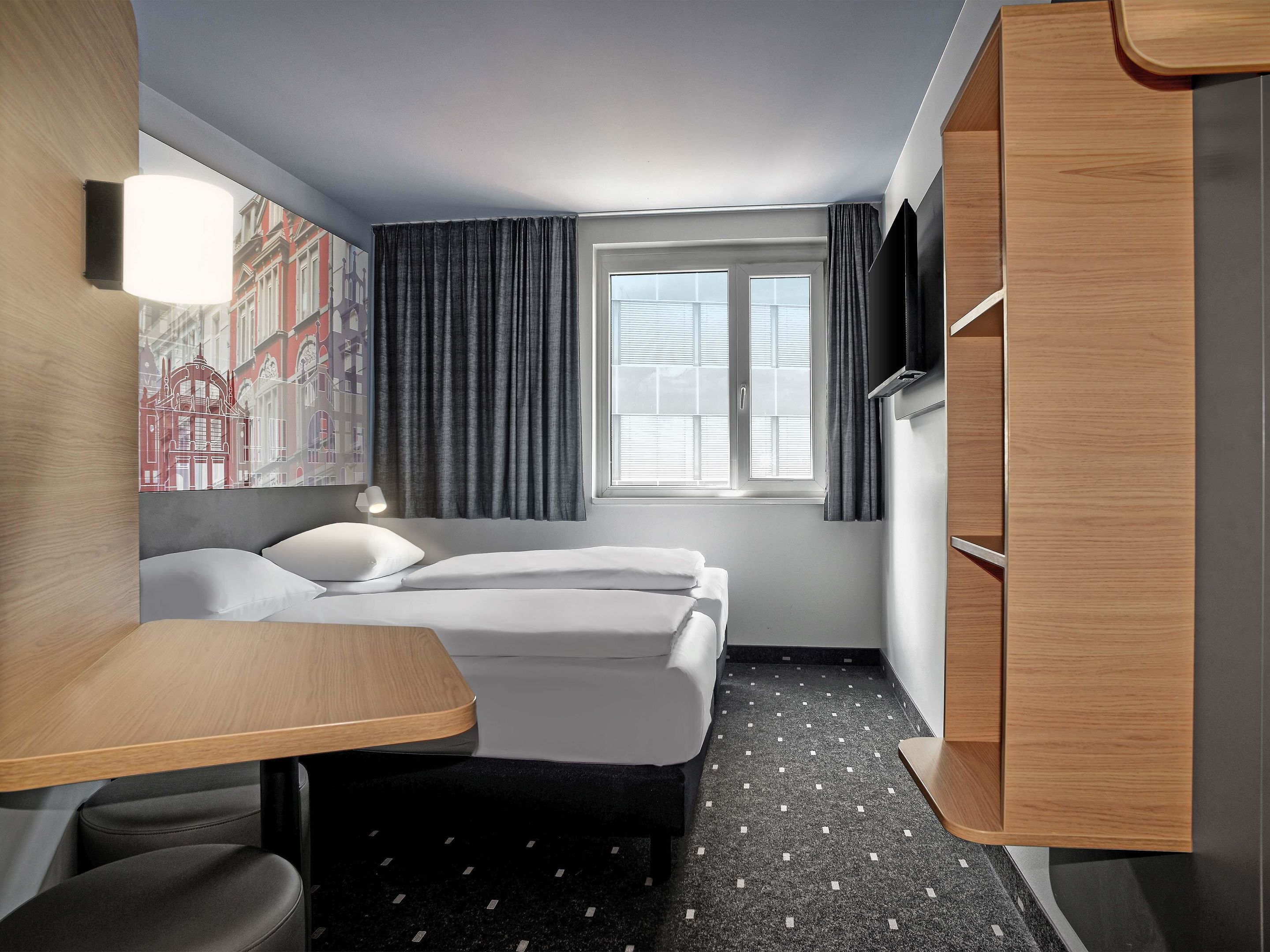Una habitación de hotel con cama, escritorio, silla, armario y ventana con cortinas.