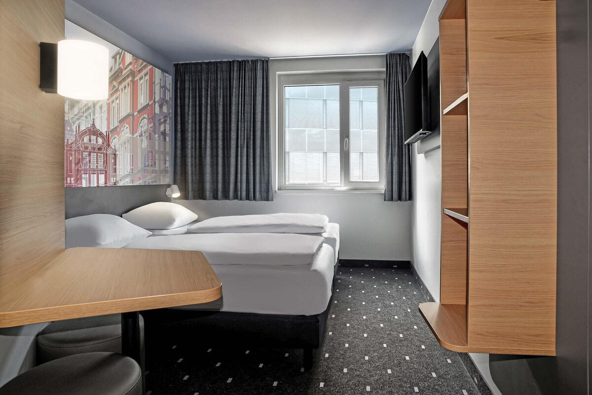 Ein Hotelzimmer mit einem Bett, einem Schreibtisch, einem Stuhl, einem Kleiderschrank und einem Fenster mit Vorhängen.