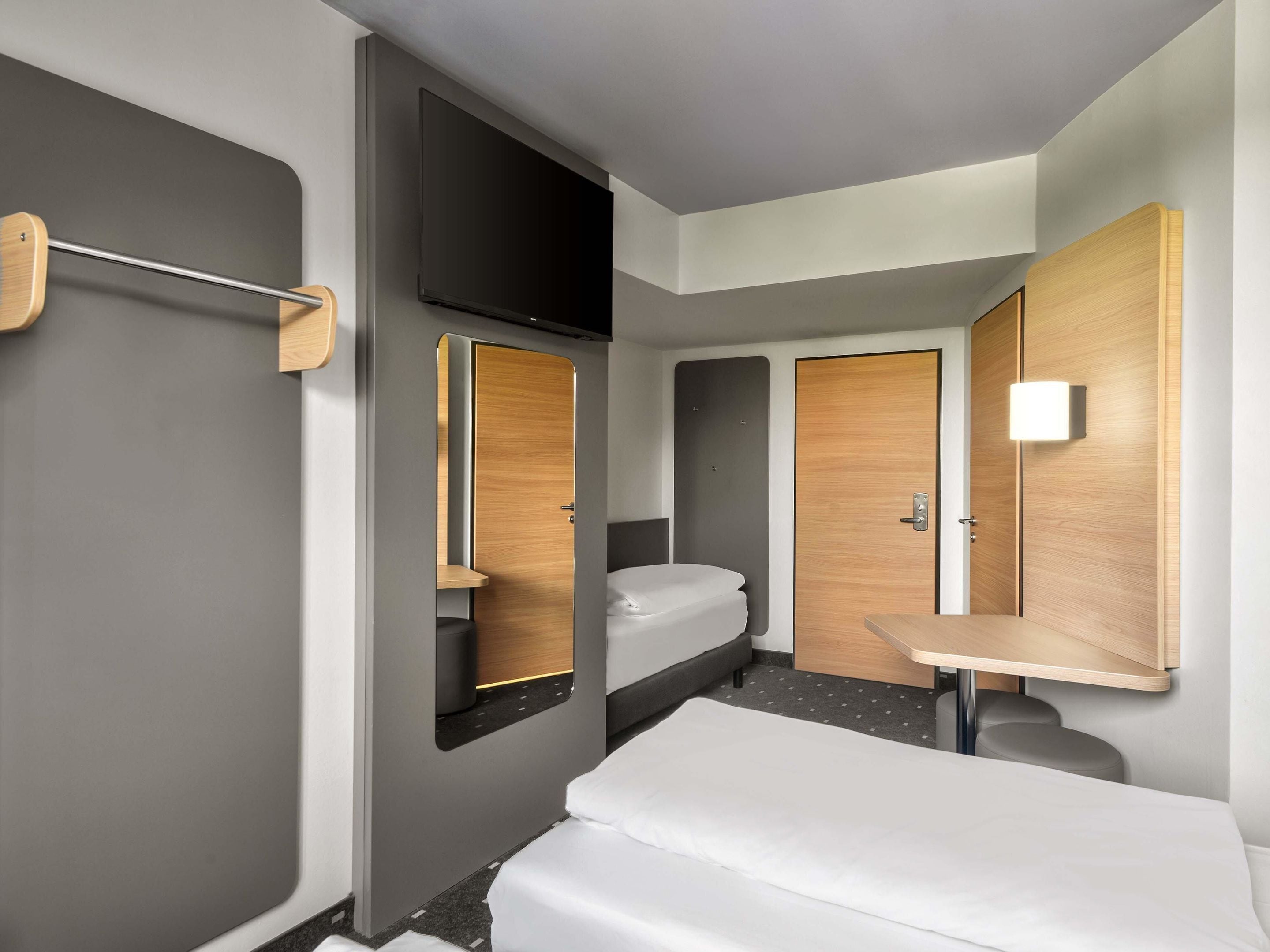 A compact hotel room with a bed, a small bathroom with a toilet and sink, a flat-screen TV mounted on the wall, and a wooden-framed mirror.