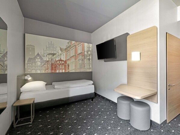 A modern hotel room with a bed, a small bedside table, a mounted TV, and a large window with a cityscape view.