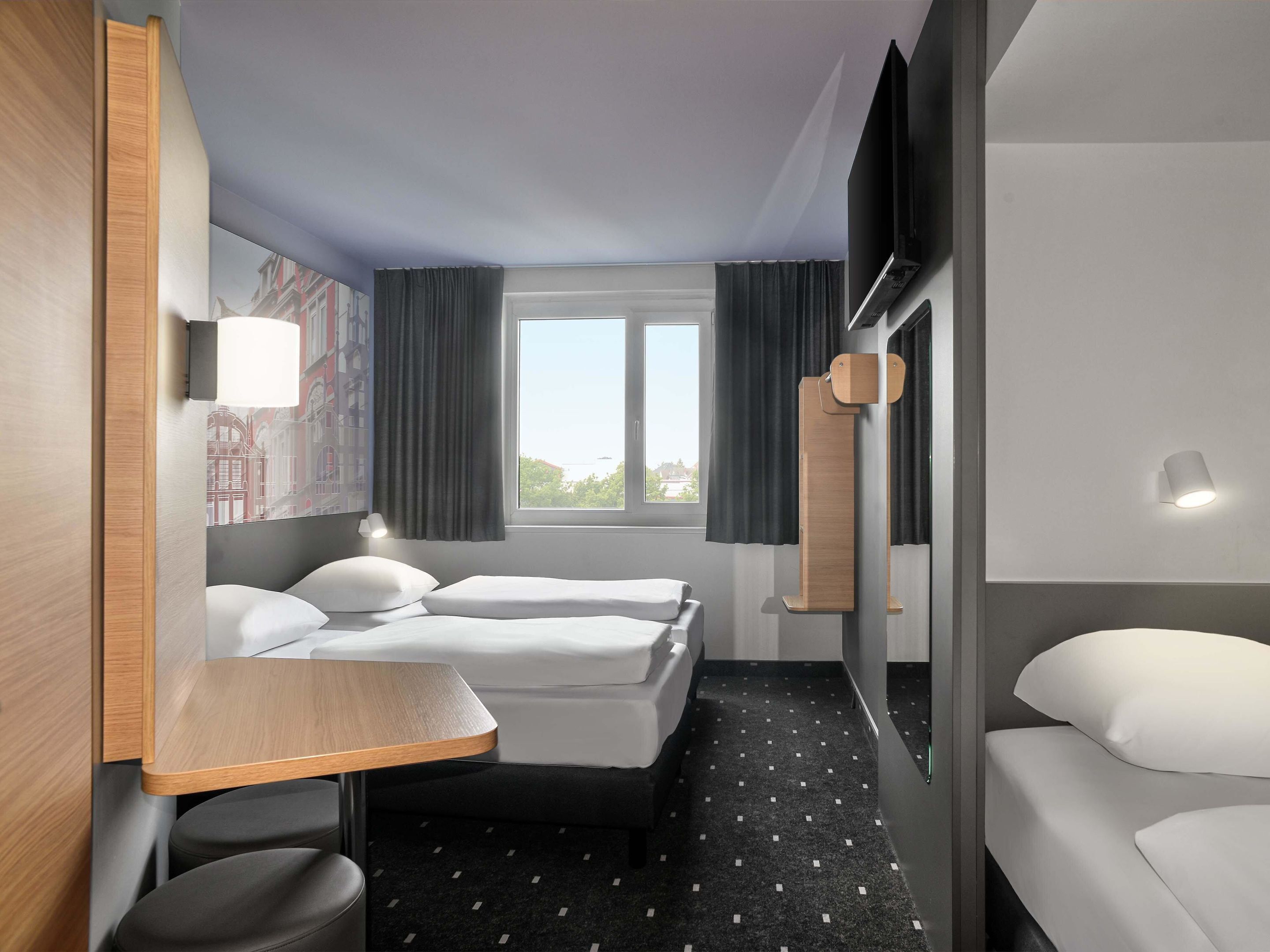 A hotel room with two beds, a small table, a chair, a window with curtains, and a wall-mounted TV.