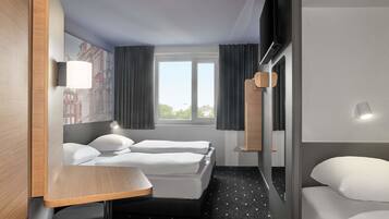 A hotel room with two beds, a small table, a chair, a window with curtains, and a wall-mounted TV.
