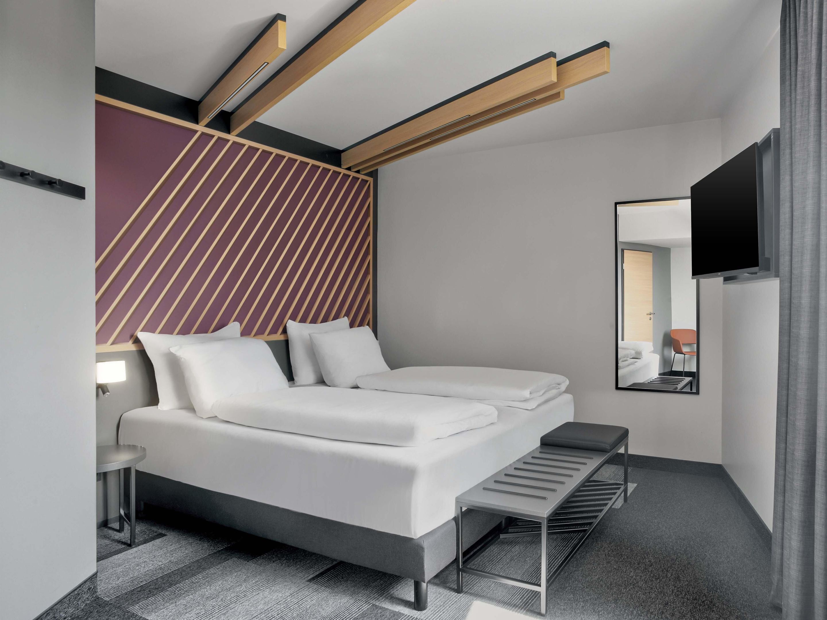 A modern hotel room with a bed, a small table, a bench, a mirror, and a TV.