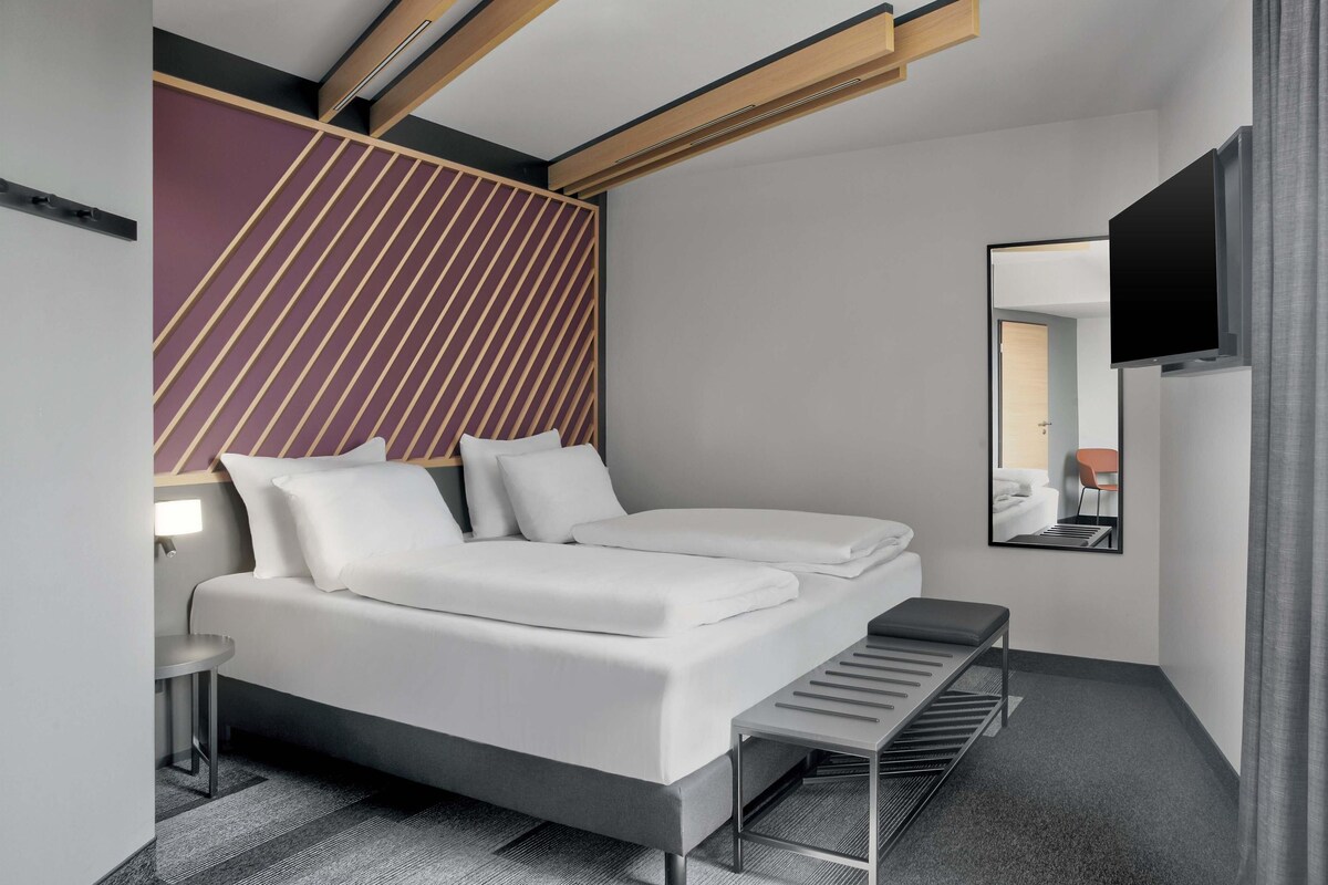Ein modernes Hotelzimmer mit einem Bett, einem kleinen Tisch, einer Bank, einem Spiegel und einem Fernseher.