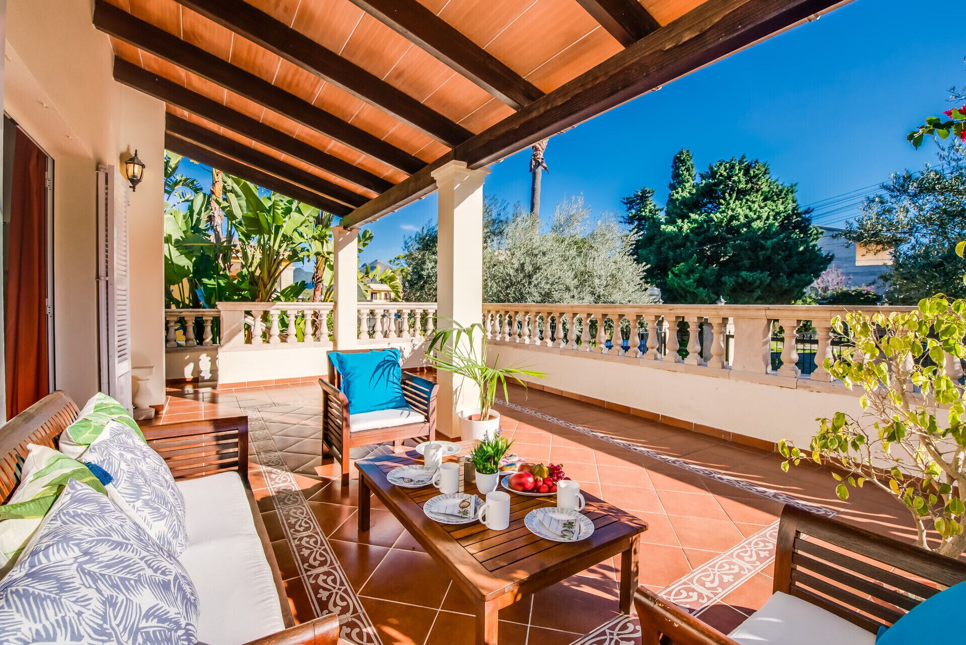 A covered patio with a wooden table set for dining, a sofa with cushions, and a potted plant.