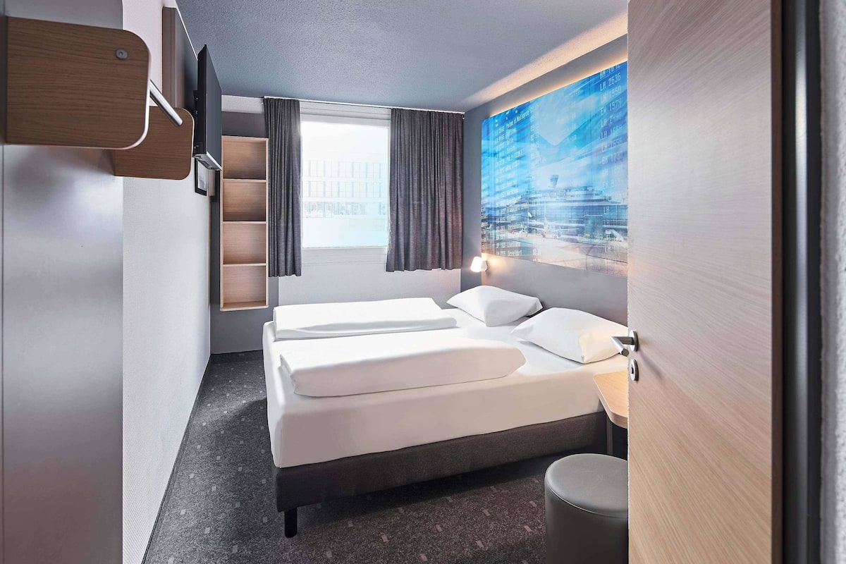 A modern hotel room with a large bed, a small bedside table, a wall-mounted TV, and a window with curtains.