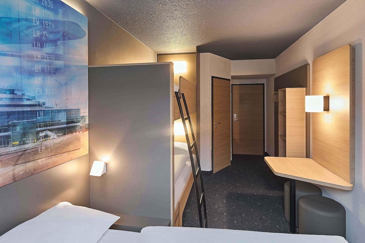 A hotel room with a bed, a desk, a chair, a ladder, and a poster on the wall.