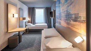 A modern hotel room with a large bed, a desk, a chair, a small table, a mirror, and a window with curtains. - B&B Hotel Köln-Airport (Cologne)