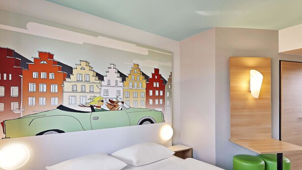 Habitación de hotel con cama, reposapiés verde y un mural en la pared con un coche y edificios.