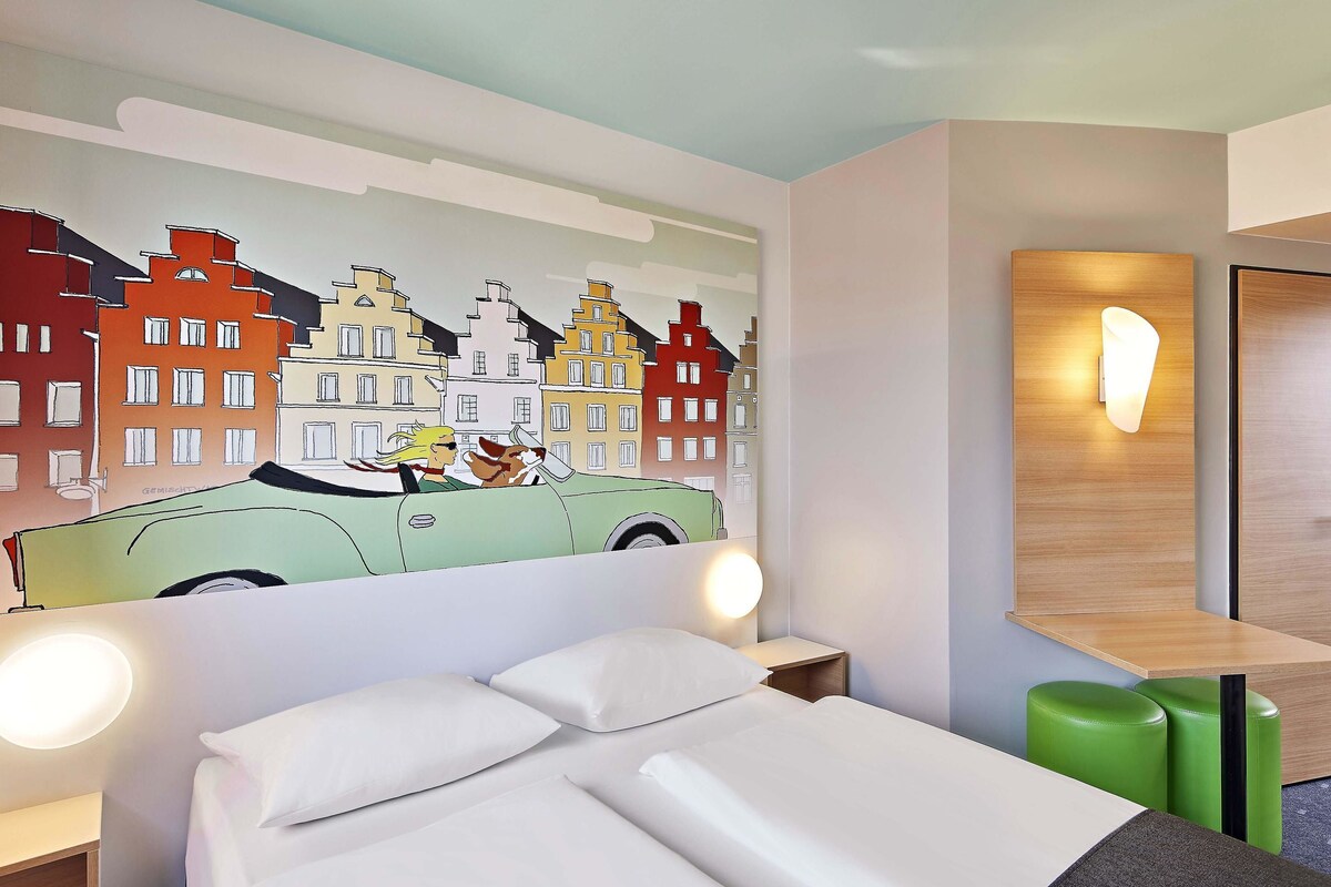 Hotellihuone, jossa on sänky, vihreä rahi ja seinämaalaus, jossa on kuvattuna auto ja rakennuksia.