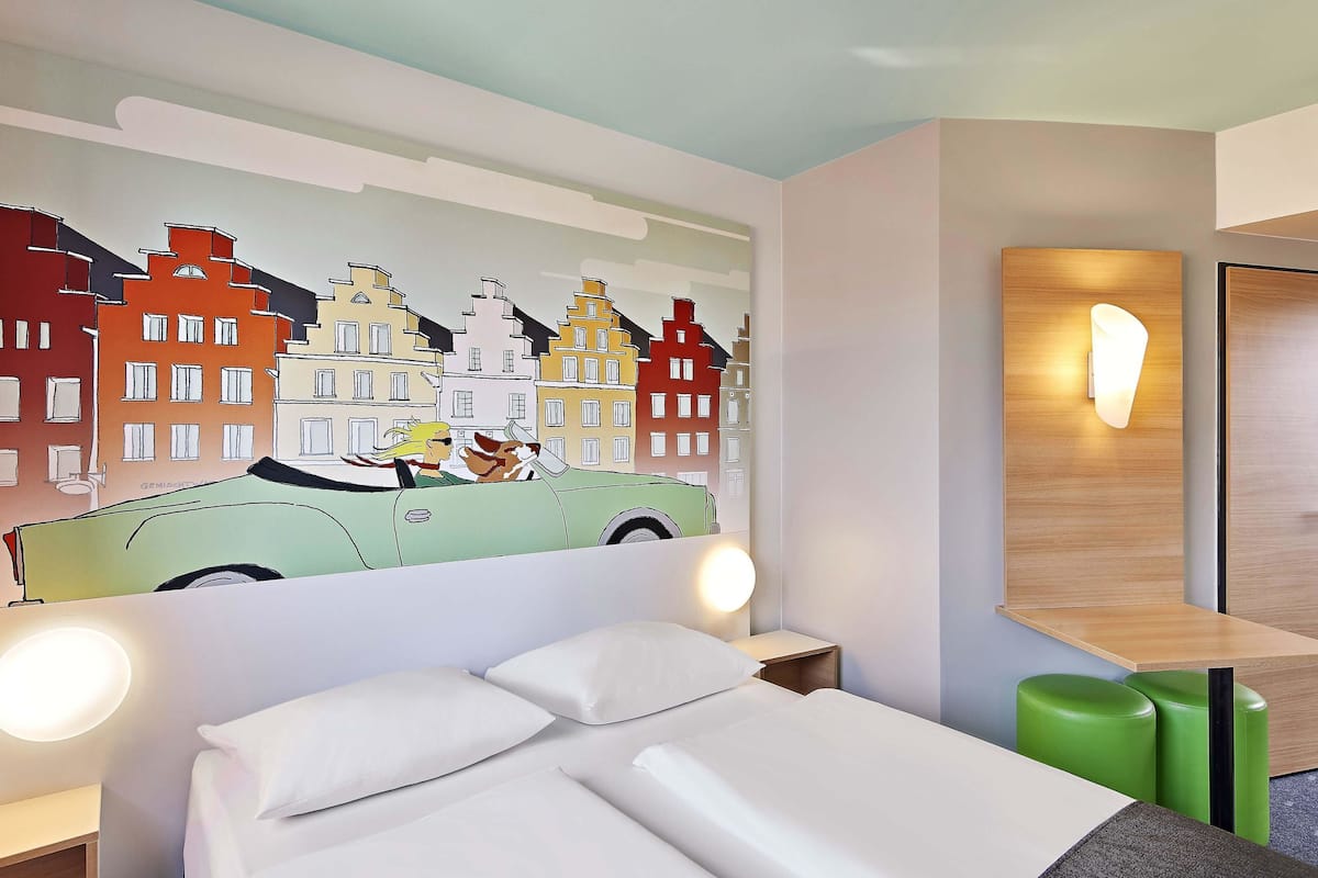 Une chambre d’hôtel avec un lit, un pouf vert et une fresque murale représentant une voiture et des bâtiments.