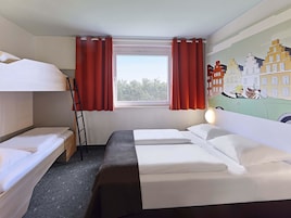 Habitación de hotel con literas, una ventana con cortinas rojas, una lámpara y un mural con una escena urbana.