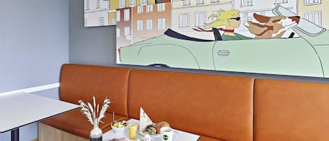 A modern hotel room with a brown leather sofa, a white table with breakfast items, and a cityscape painting on the wall.