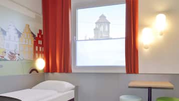 Una stanza con un letto, un tavolino, uno sgabello verde, una lampada a parete e una finestra vista su una torre.