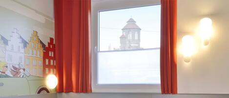 A room with a bed, a small table, a green stool, a wall-mounted light, and a window with a view of a tower.