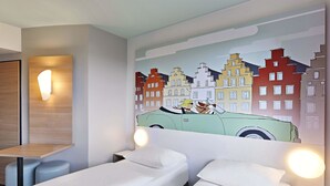 Habitación de hotel con dos camas individuales, una mesita de noche de madera, una lámpara de pared y un mural de un paisaje urbano colorido.