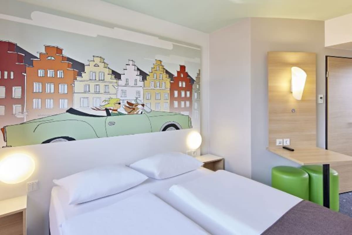 A hotel room with a bed, a wall mural of a cityscape, a desk with a lamp, and a green stool.