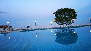 Piscina con sillones de descanso y sombrillas, rodeada de árboles y un edificio.