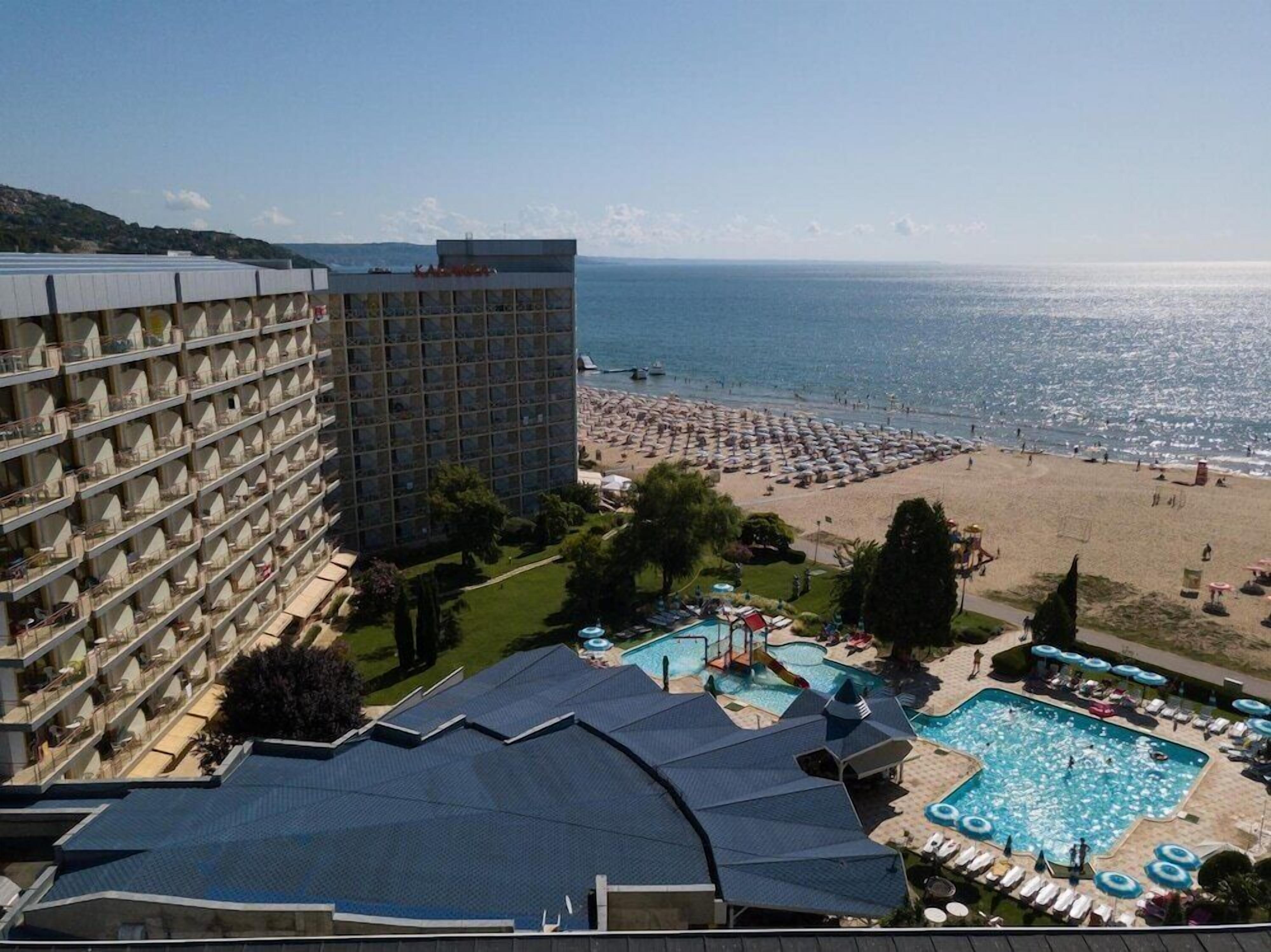 A hotel with multiple floors, a pool area with lounge chairs and umbrellas, and a beach with umbrellas and people.