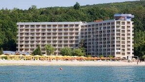 Un hotel de varios pisos con balcones, situado cerca de una playa con sombrillas y gente.