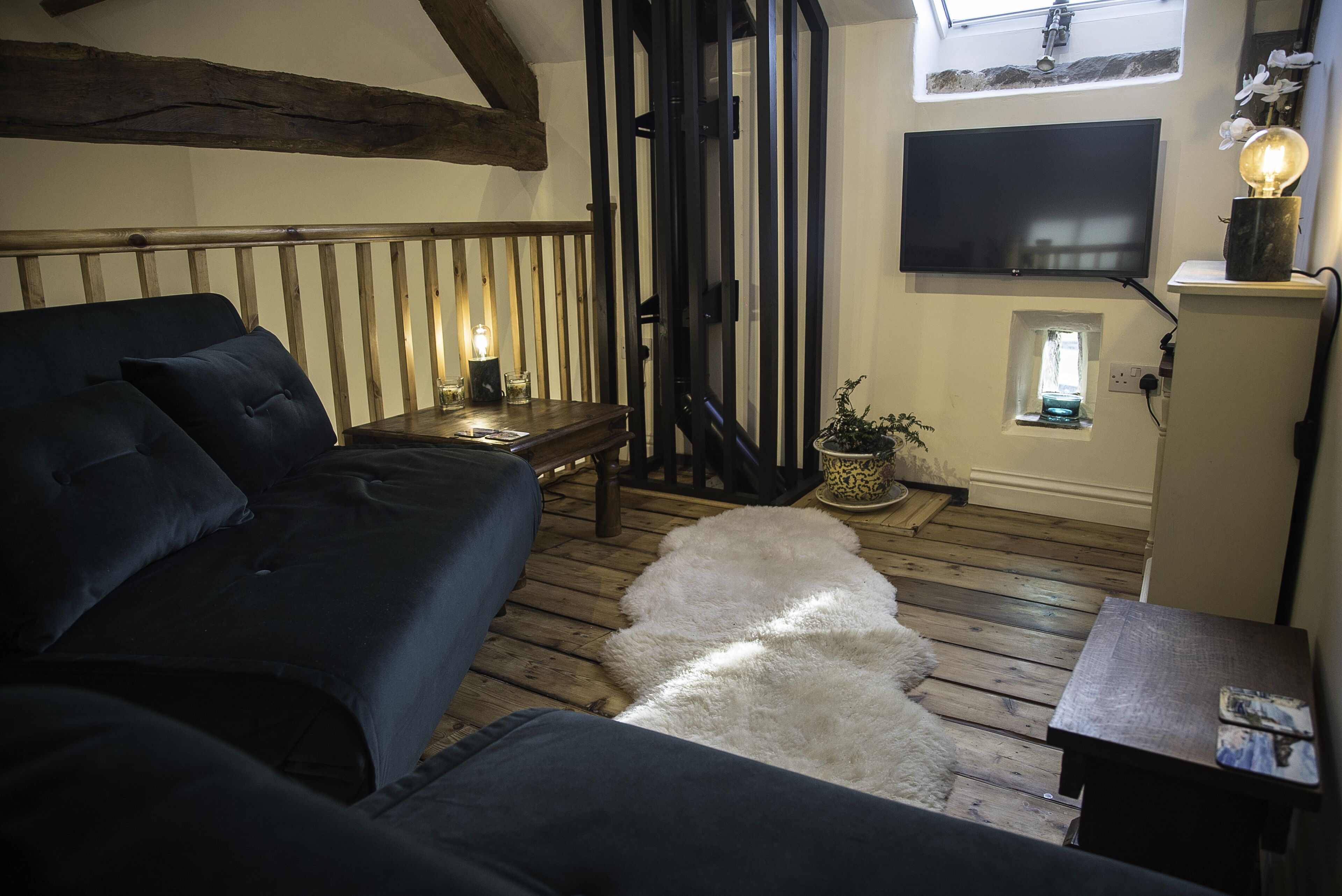 A cozy room with a dark blue sofa, a wooden floor, a white furry rug, a small table with a lamp, and a TV mounted on the wall.
