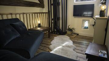 A cozy room with a dark blue sofa, a wooden floor, a white furry rug, a small table with a lamp, and a TV mounted on the wall.