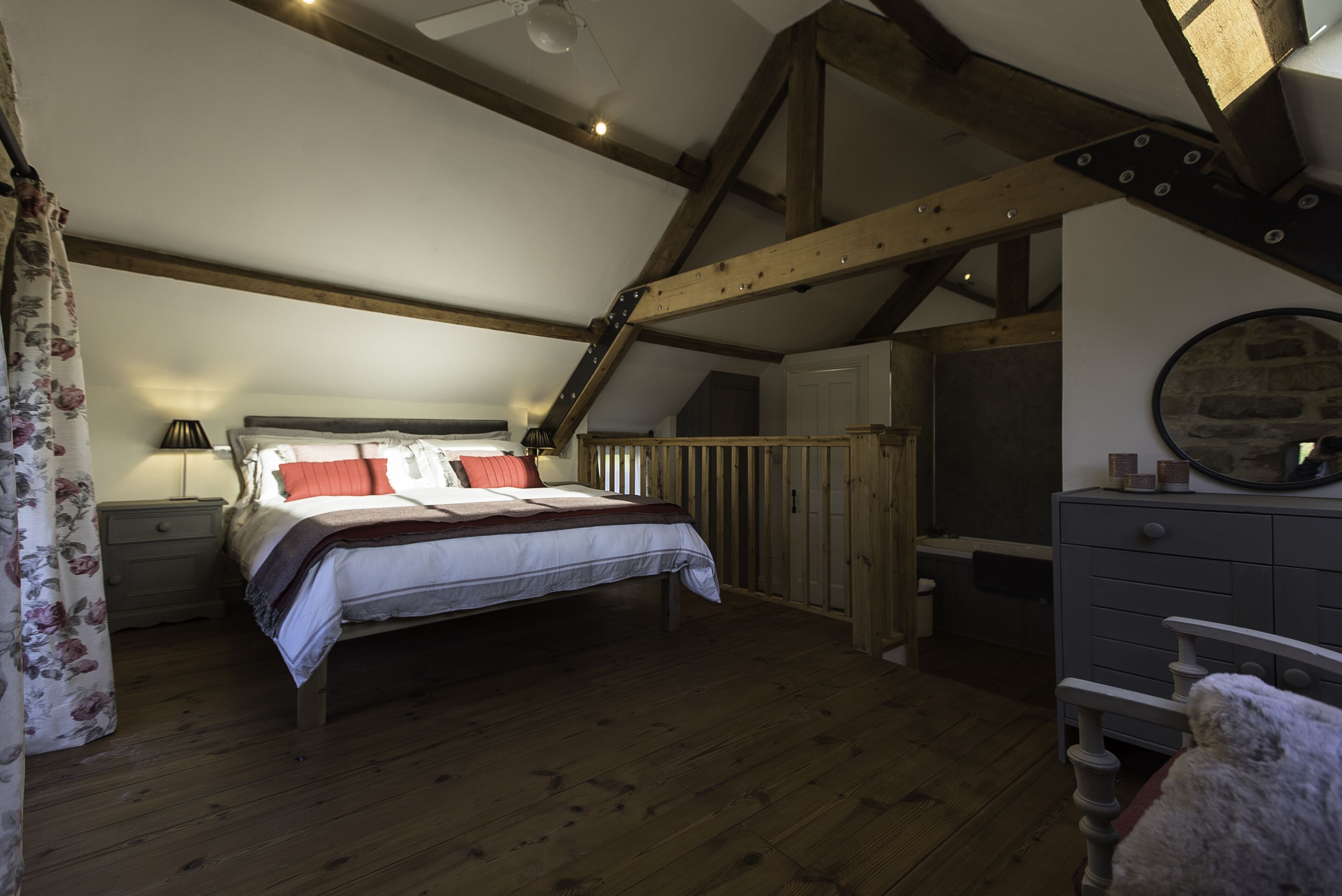 Una camera da letto con pavimento in legno, un letto con biancheria rossa e bianca, un comodino, una cassettiera e uno specchio appeso al muro.