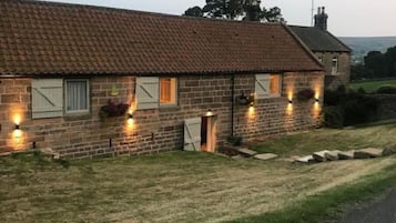 Una cabaña de piedra con techo de tejas, varias ventanas con contraventanas e iluminación exterior.