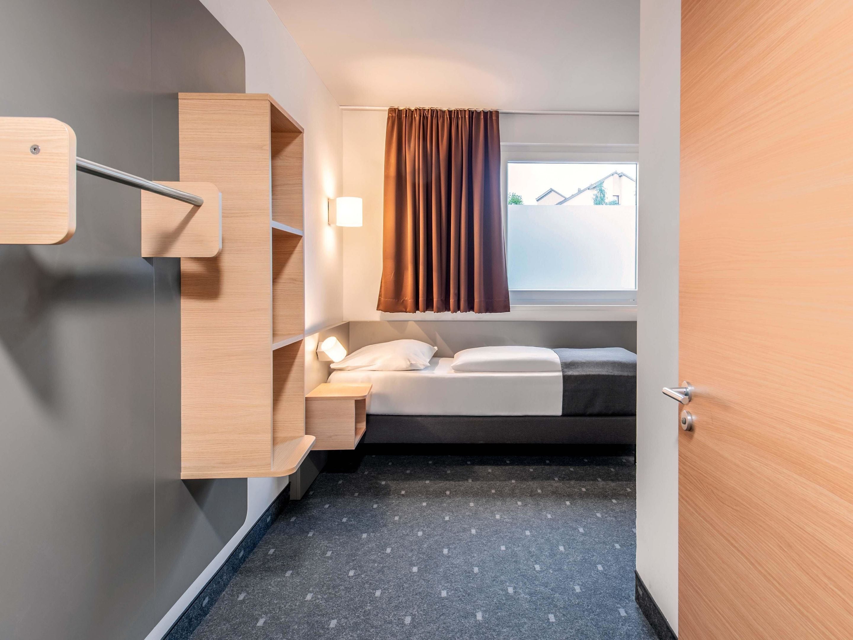 A compact hotel room with a bed, a bedside table, a built-in shelf, and a window with curtains.
