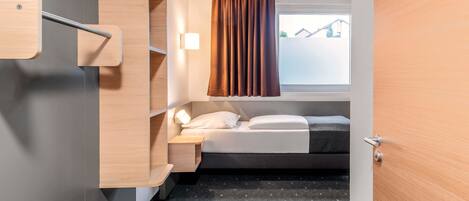 A compact hotel room with a bed, a bedside table, a built-in shelf, and a window with curtains.