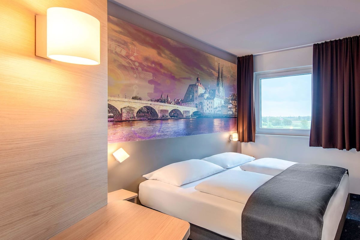 A hotel room with a bed, a desk, a wall-mounted lamp, a window with curtains, and a cityscape painting.