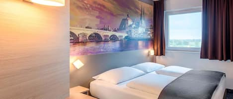 Habitación de hotel con cama, escritorio, lámpara de pared, ventana con cortinas y una pintura de paisaje urbano.