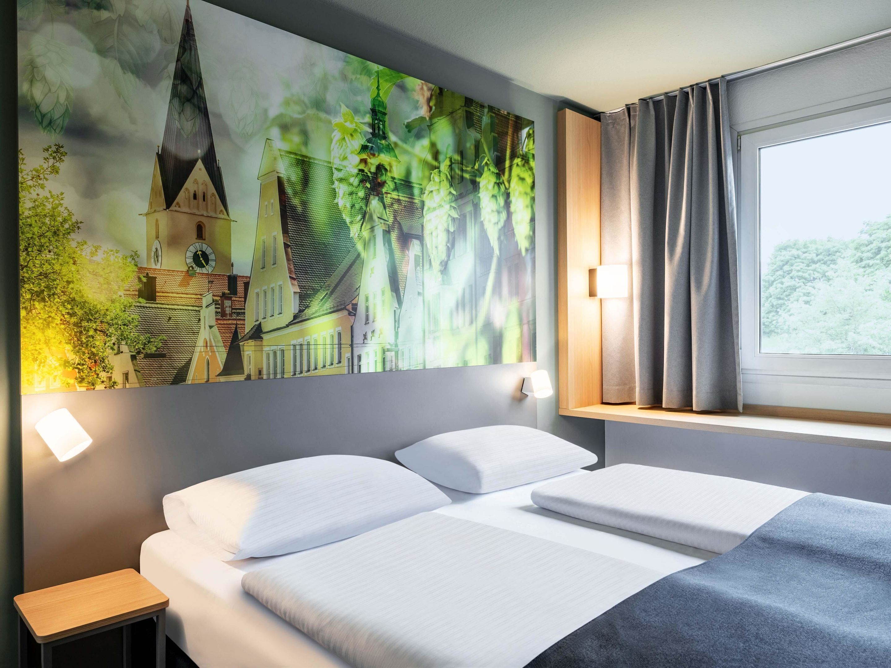 A hotel room with a bed, a nightstand, a wall-mounted lamp, a window with curtains, and a large painting of a building and greenery.