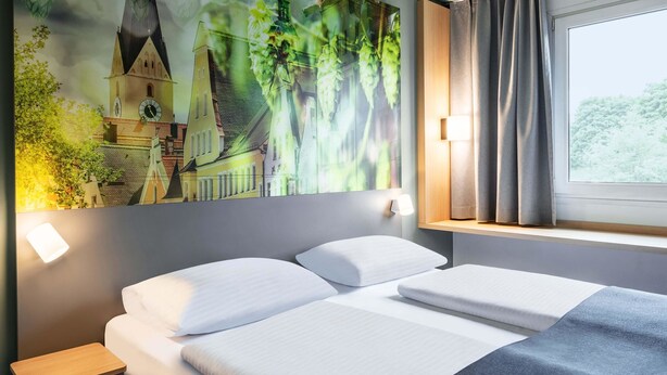 A hotel room with a bed, a nightstand, a wall-mounted lamp, a window with curtains, and a large painting of a building and greenery. - B&B HOTEL Ingolstadt-Ost (Ingolstadt)