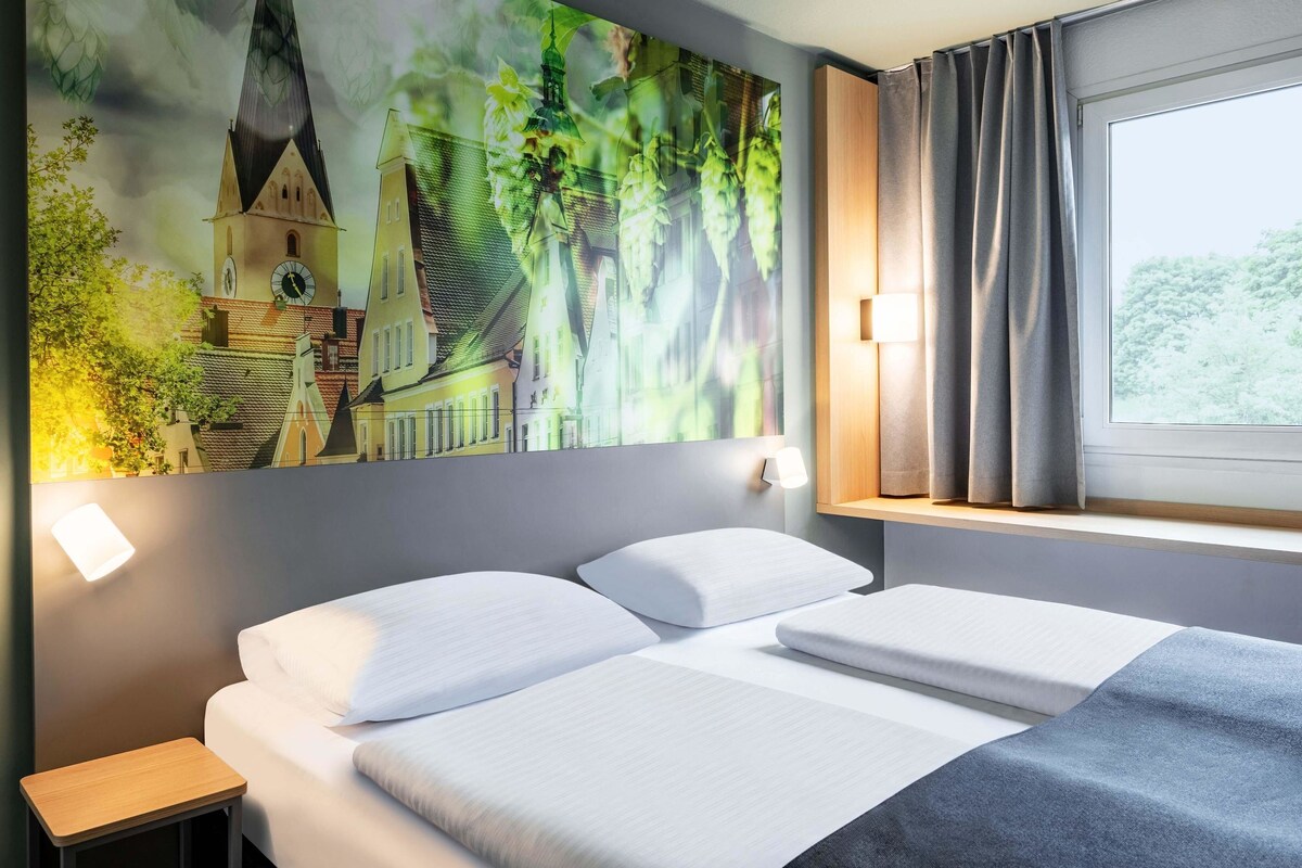 A hotel room with a bed, a nightstand, a wall-mounted lamp, a window with curtains, and a large painting of a building and greenery.