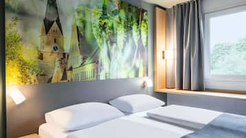 A hotel room with a bed, a nightstand, a wall-mounted lamp, a window with curtains, and a large painting of a building and greenery.