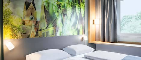 Una habitación de hotel con una cama, una mesita de noche, una lámpara de pared, una ventana con cortinas y un cuadro grande de un edificio y vegetación.