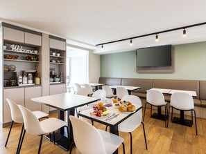 A modern dining area with white tables and chairs, a 'Welcome' sign, and a TV mounted on the wall.