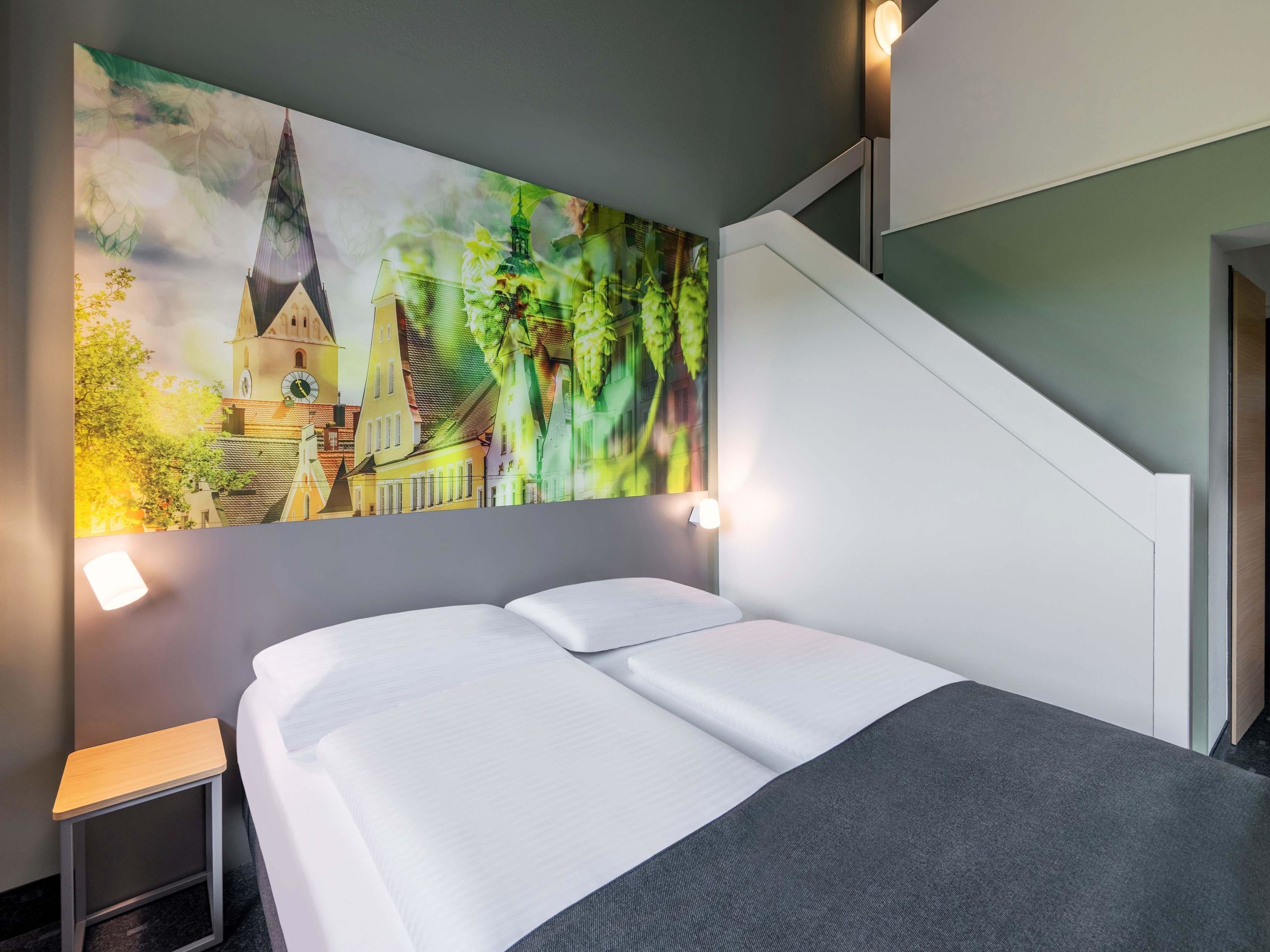 A hotel room with a bed, a small bedside table, and a large painting of a town scene on the wall.